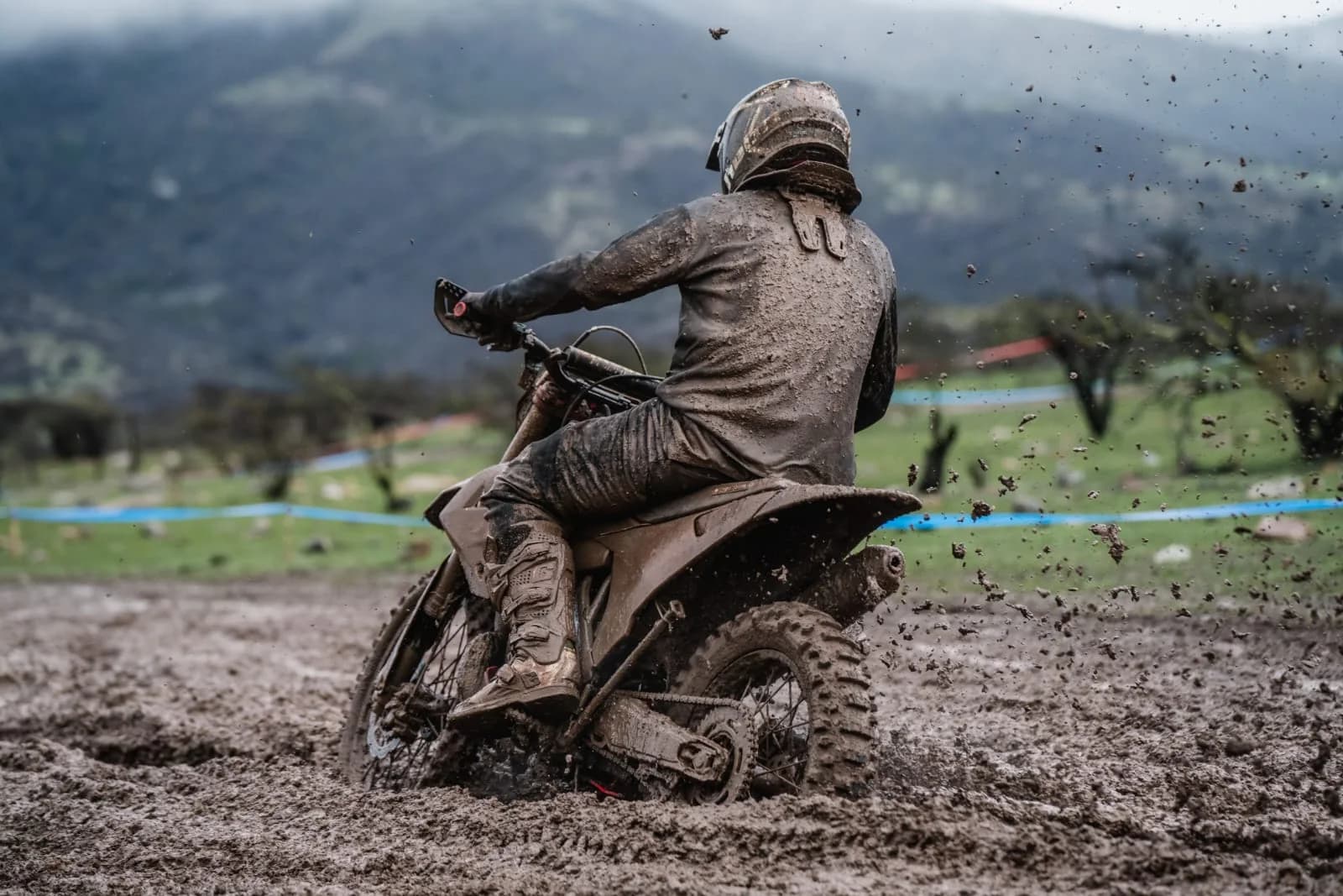 Piloto cubierto de barro acelerando su moto de enduro en un tramo inundado, con la cordillera al fondo
